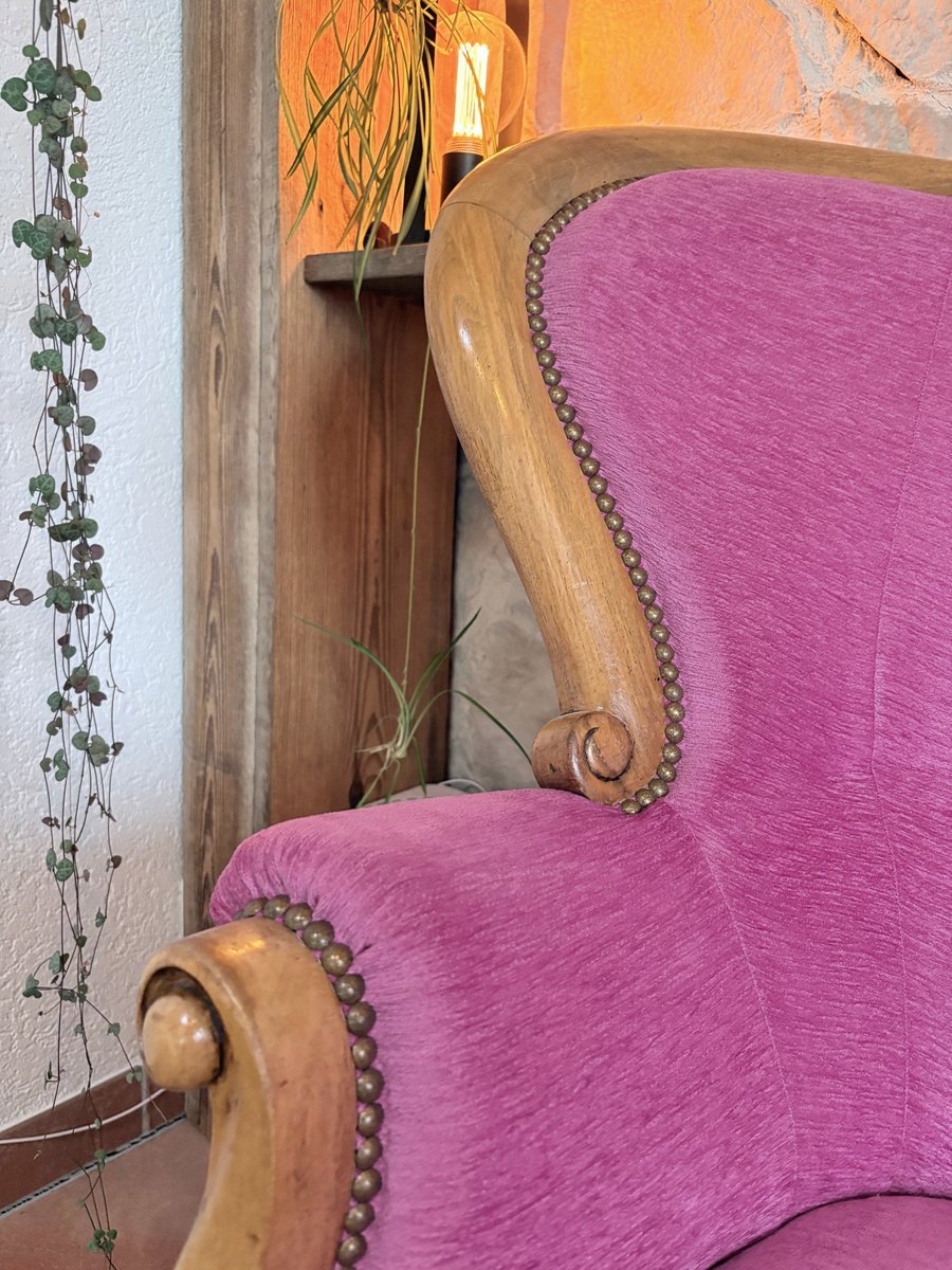 Accoudoir sculpté d’un canapé en velours framboise, lampe allumée sur le mur de pierre